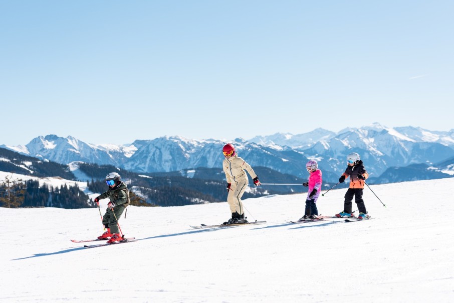 Eine Person und drei Kinder fahren bei sonnigem Wetter Ski in den Bergen von Hochkönig im Winterurlaub im Hotel Bachschmied. ©TVB Hochkönig/Miriam Lottes
