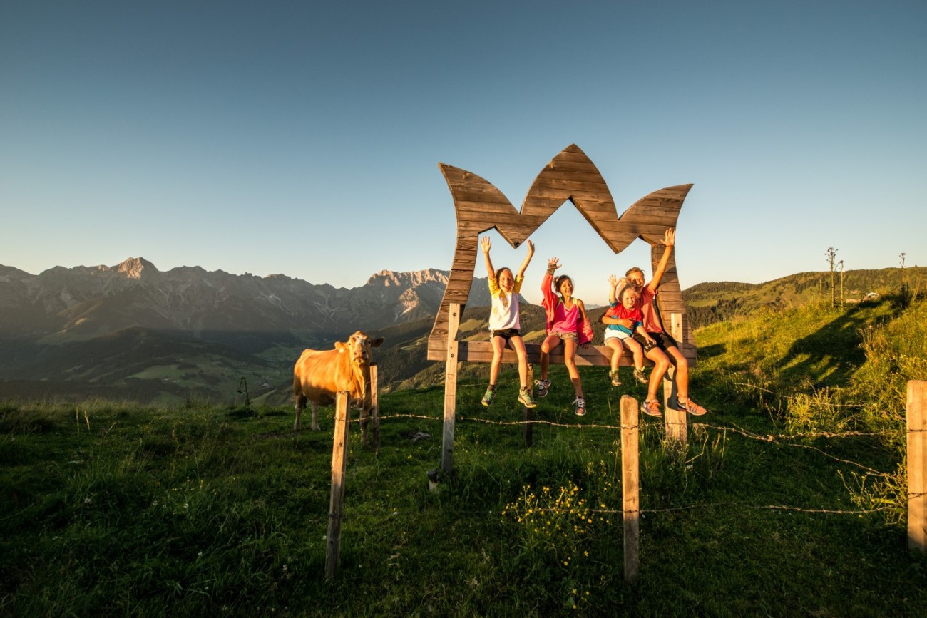 Kinder sitzen fröhlich auf einem Holzrahmen in den Bergen neben einer Kuh, Sommerurlaub am Hochkönig. ©TVB_Hochkönig/Christian Schartner