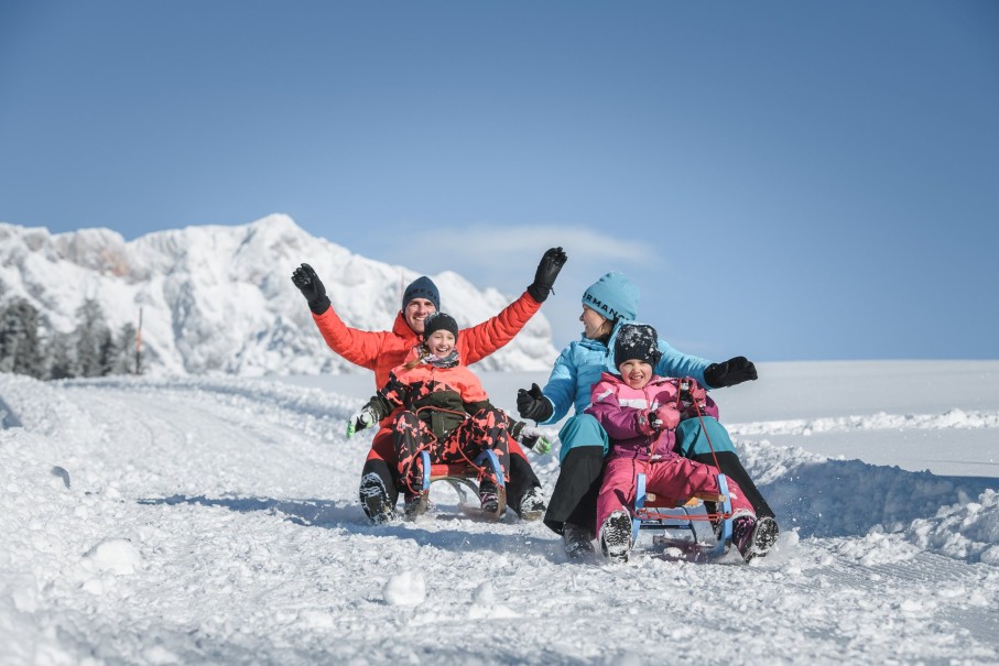 Menschen rodeln fröhlich im Schnee vor einer malerischen Berglandschaft am Hochkönig im Winterurlaub im Hotel Bachschmied. ©TVB Hochkönig