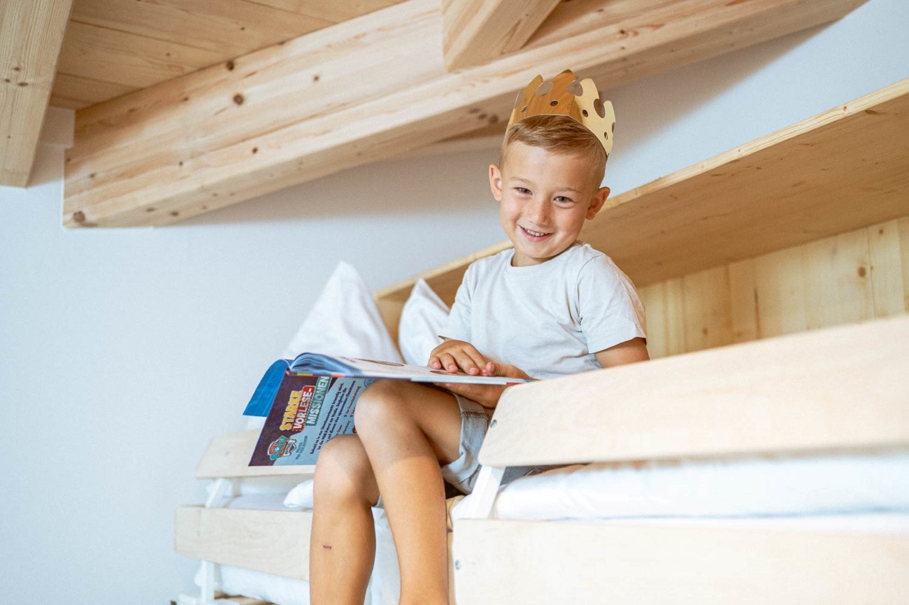 Ein lächelnder Junge mit Krone liest ein Buch auf einem Hochbett im Familienurlaub im Hotel Bachschmied.